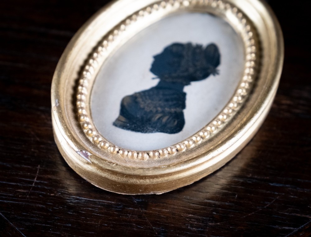 An oval framed Regency-era miniature silhouette depicting the profile of a lady embellished with fine gold detailing, set within a beaded gilt frame. View VI