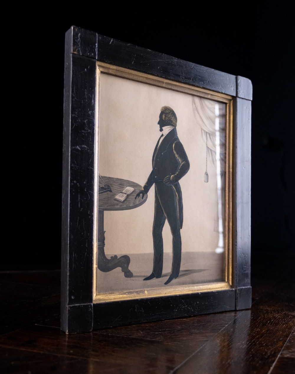 An early nineteenth-century full-length silhouette portrait of a standing gentleman in profile beside a pedestal table, presented in a black rectangular frame with a gilded inner border. View VI