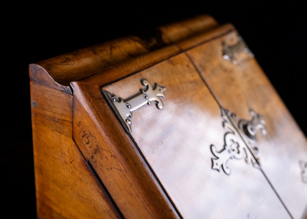 A Victorian walnut stationery cabinet featuring a sloped front with double doors, brass strapwork hinges, and a lower drawer. View VI