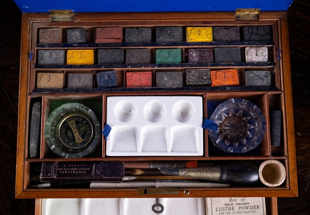 A Victorian mahogany artist watercolour box featuring an open lid lined with blue gilt-stamped paper, fitted compartments containing colour cakes, glass dishes, and a lower drawer holding a ceramic palette. View V