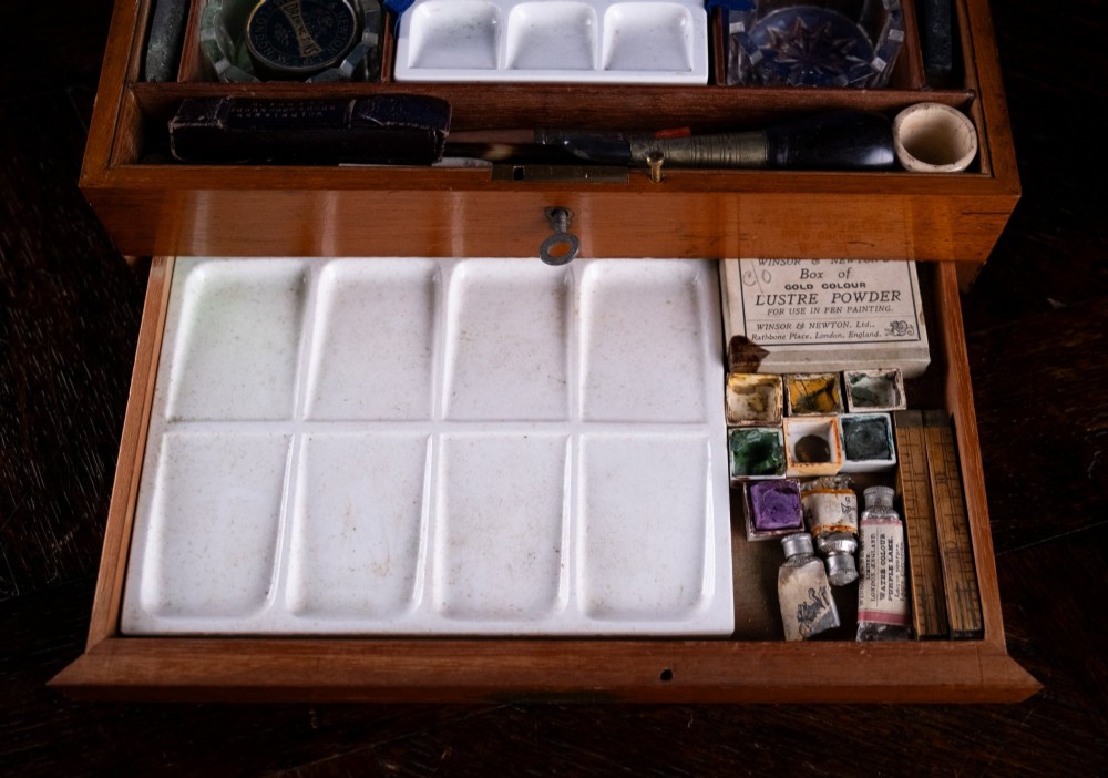 A Victorian mahogany artist watercolour box featuring an open lid lined with blue gilt-stamped paper, fitted compartments containing colour cakes, glass dishes, and a lower drawer holding a ceramic palette. View VI