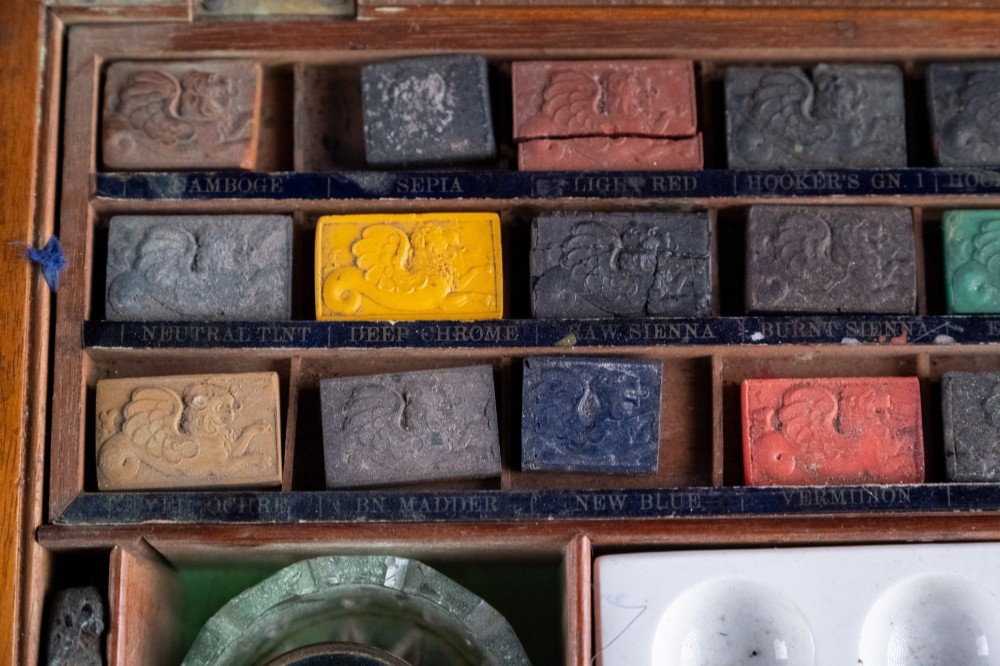 A Victorian mahogany artist watercolour box featuring an open lid lined with blue gilt-stamped paper, fitted compartments containing colour cakes, glass dishes, and a lower drawer holding a ceramic palette. View VII