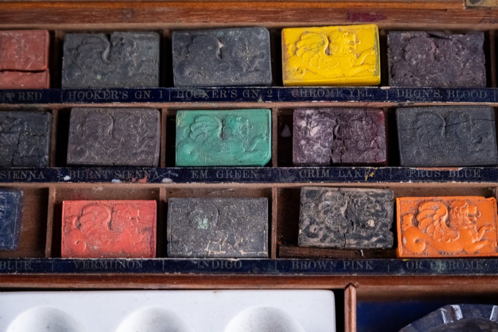 A Victorian mahogany artist watercolour box featuring an open lid lined with blue gilt-stamped paper, fitted compartments containing colour cakes, glass dishes, and a lower drawer holding a ceramic palette. View VIII