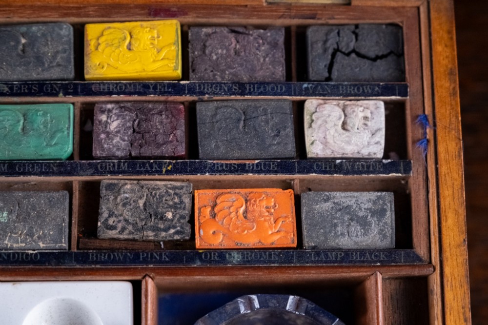 A Victorian mahogany artist watercolour box featuring an open lid lined with blue gilt-stamped paper, fitted compartments containing colour cakes, glass dishes, and a lower drawer holding a ceramic palette. View IX