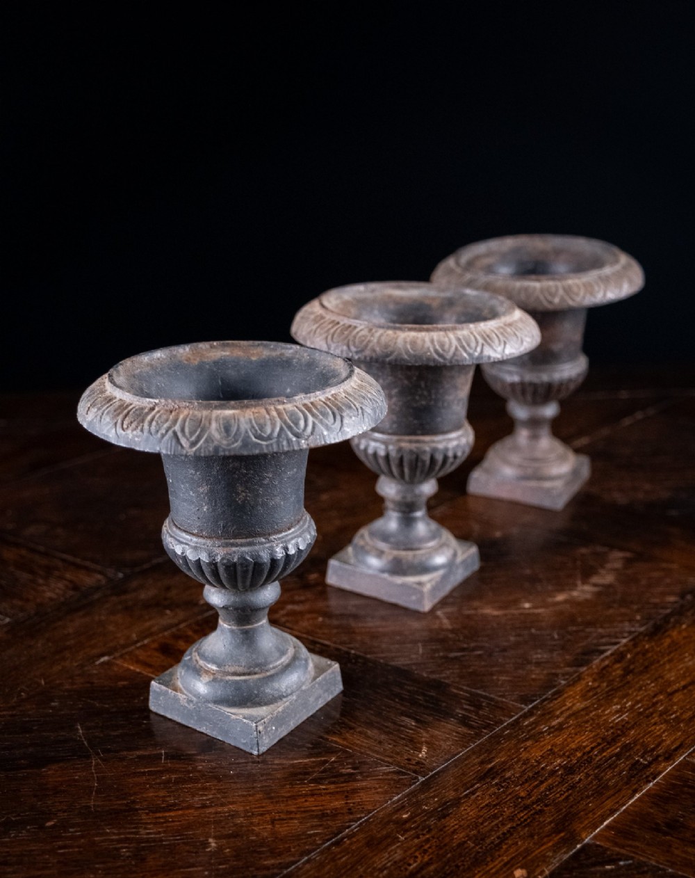 A set of three miniature cast iron urns featuring fluted campana bodies and square pedestal bases with a dark patinated finish. View VII