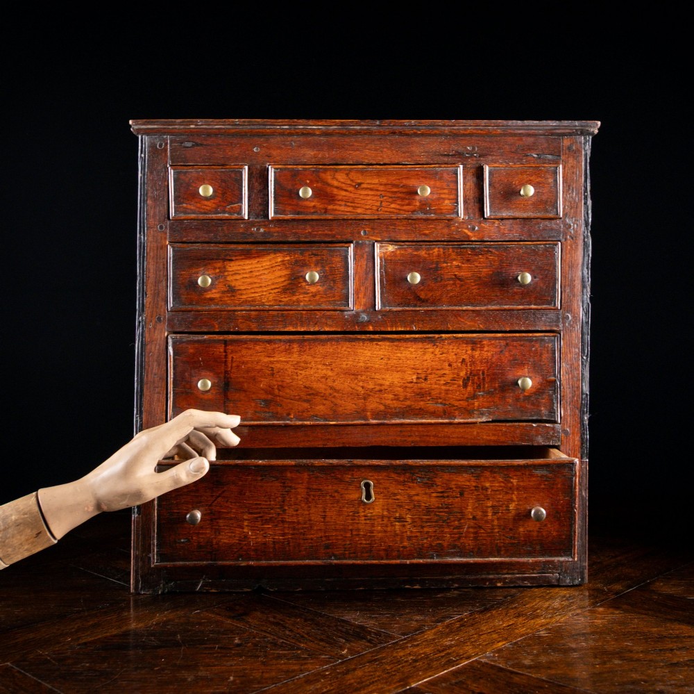 An 18th-century dark oak tabletop chest of drawers featuring seven multi-sized drawers with brass knob handles and a brass keyhole escutcheon.