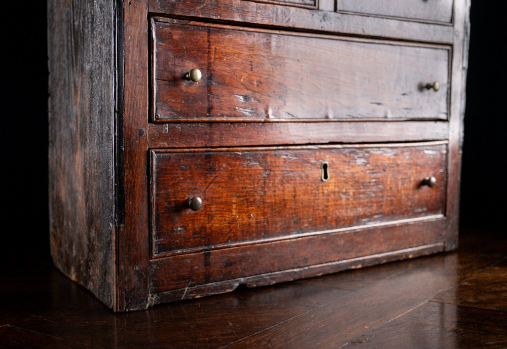 An 18th-century dark oak tabletop chest of drawers featuring seven multi-sized drawers with brass knob handles and a brass keyhole escutcheon. View V