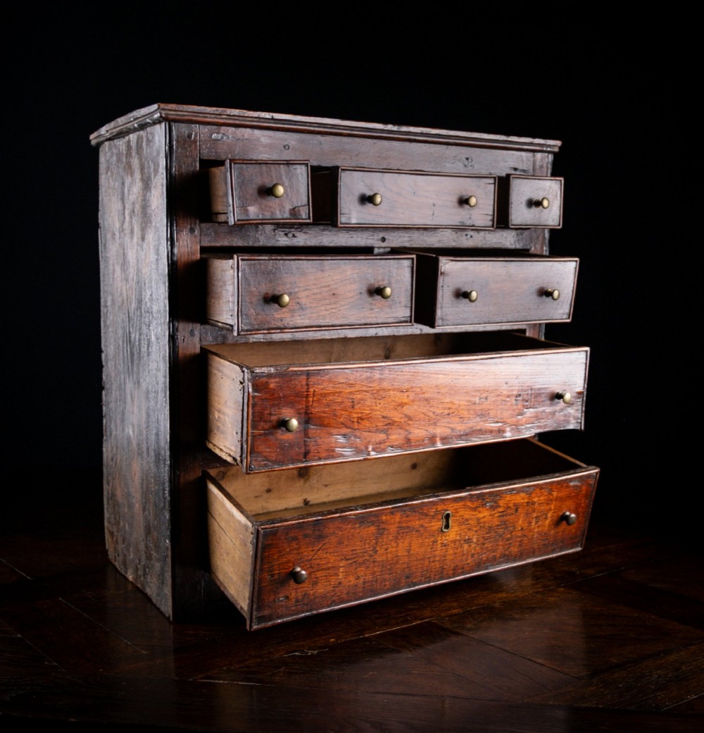 An 18th-century dark oak tabletop chest of drawers featuring seven multi-sized drawers with brass knob handles and a brass keyhole escutcheon. View VI