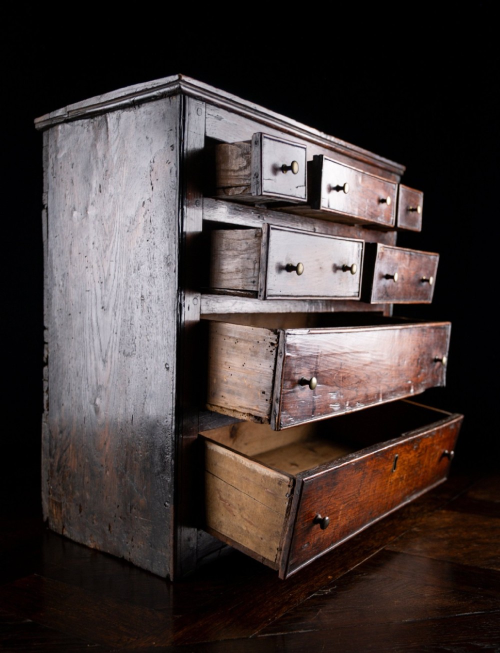 An 18th-century dark oak tabletop chest of drawers featuring seven multi-sized drawers with brass knob handles and a brass keyhole escutcheon. View VII