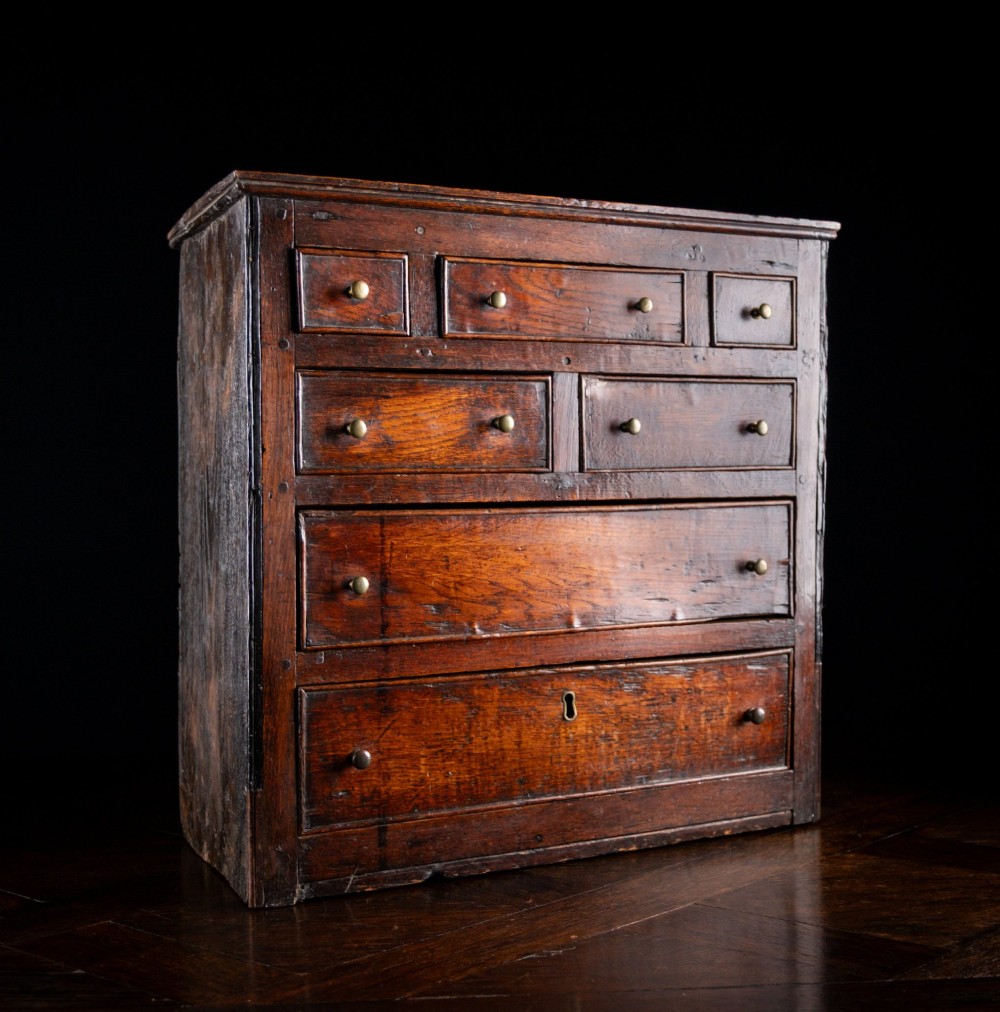 An 18th-century dark oak tabletop chest of drawers featuring seven multi-sized drawers with brass knob handles and a brass keyhole escutcheon. View IX