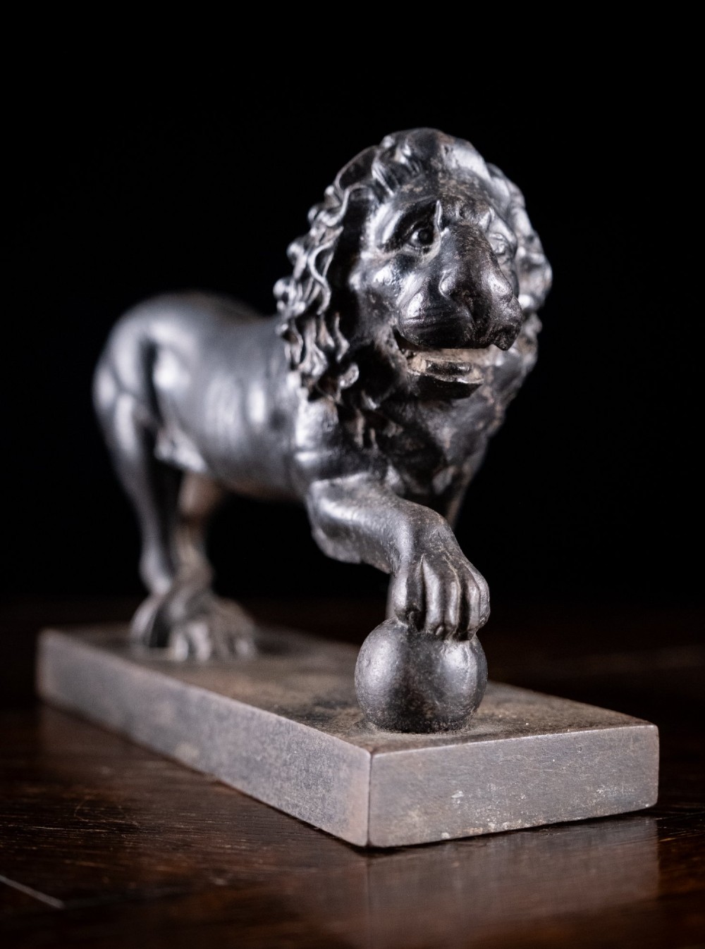 A dark cast iron lion sculpture standing on a rectangular base with one front paw resting on a sphere. View III