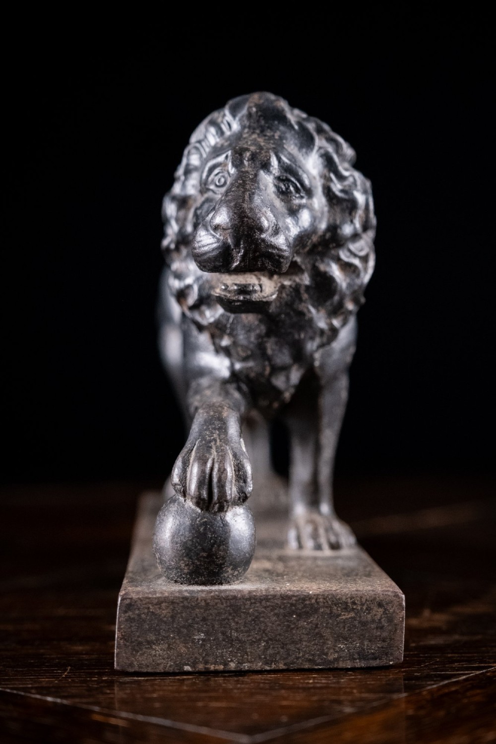 A dark cast iron lion sculpture standing on a rectangular base with one front paw resting on a sphere. View IV