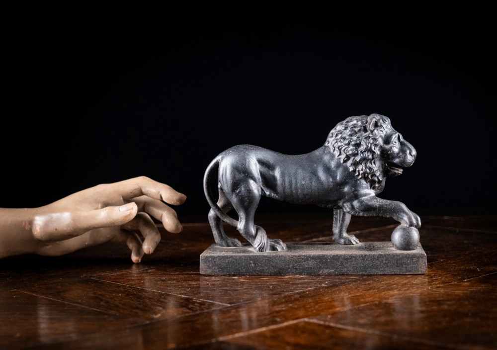 A dark cast iron lion sculpture standing on a rectangular base with one front paw resting on a sphere. View X