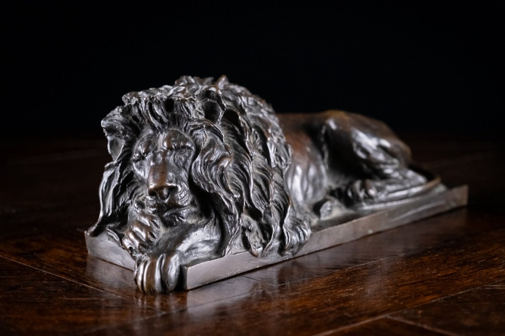 A dark brown patinated bronze sculpture depicting a recumbent sleeping lion resting its head on an integrated rectangular base.