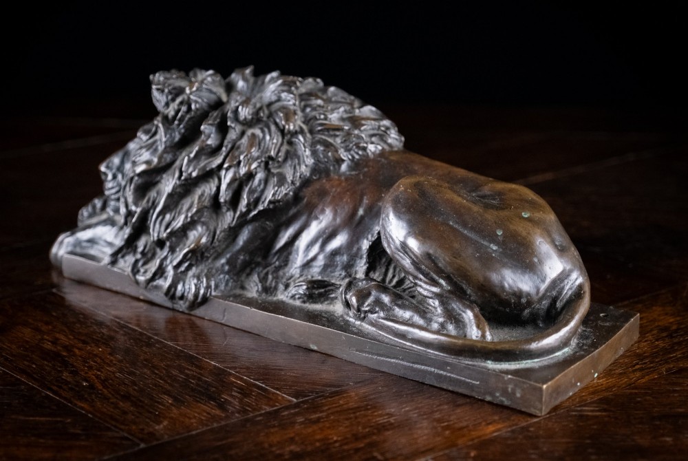 A dark brown patinated bronze sculpture depicting a recumbent sleeping lion resting its head on an integrated rectangular base. View VI