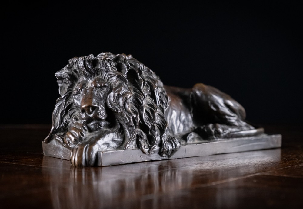 A dark brown patinated bronze sculpture depicting a recumbent sleeping lion resting its head on an integrated rectangular base. View IX