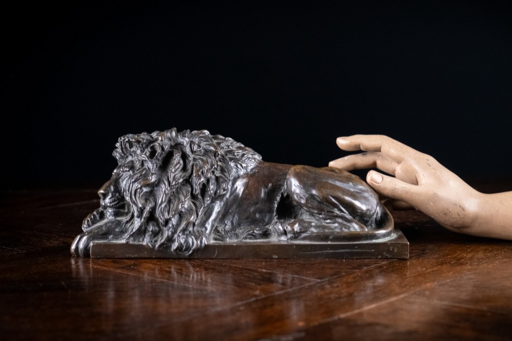 A dark brown patinated bronze sculpture depicting a recumbent sleeping lion resting its head on an integrated rectangular base. View X