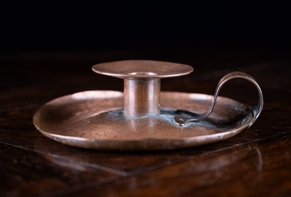 A copper Arts and Crafts chamberstick with a wide dish base, cylindrical candle sconce, and a looped metal handle.