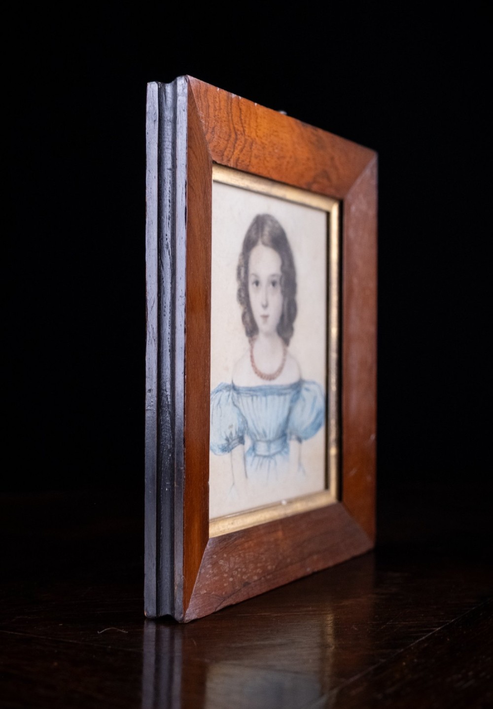 An early 19th-century watercolour portrait miniature depicting a young girl with dark curls in a blue dress, displayed in a dark wood frame. View III