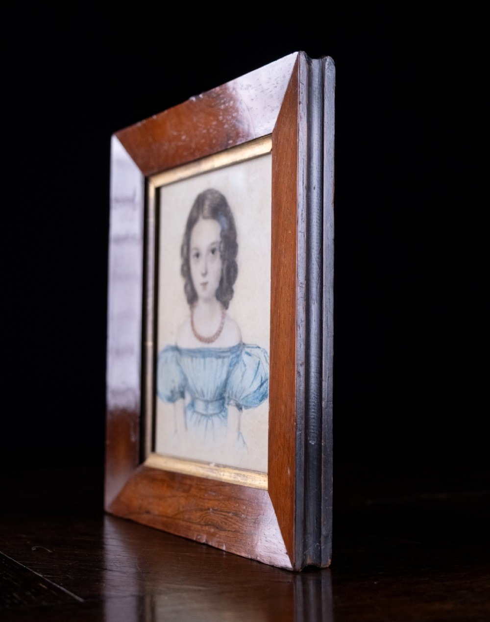 An early 19th-century watercolour portrait miniature depicting a young girl with dark curls in a blue dress, displayed in a dark wood frame. View IV