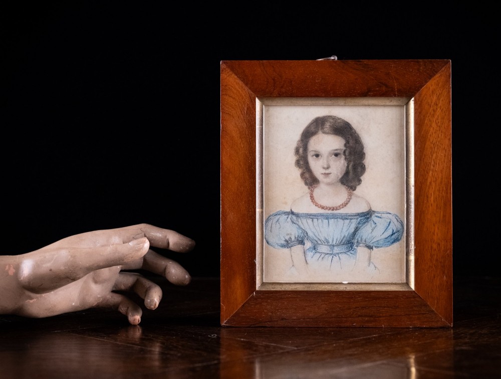 An early 19th-century watercolour portrait miniature depicting a young girl with dark curls in a blue dress, displayed in a dark wood frame. View V