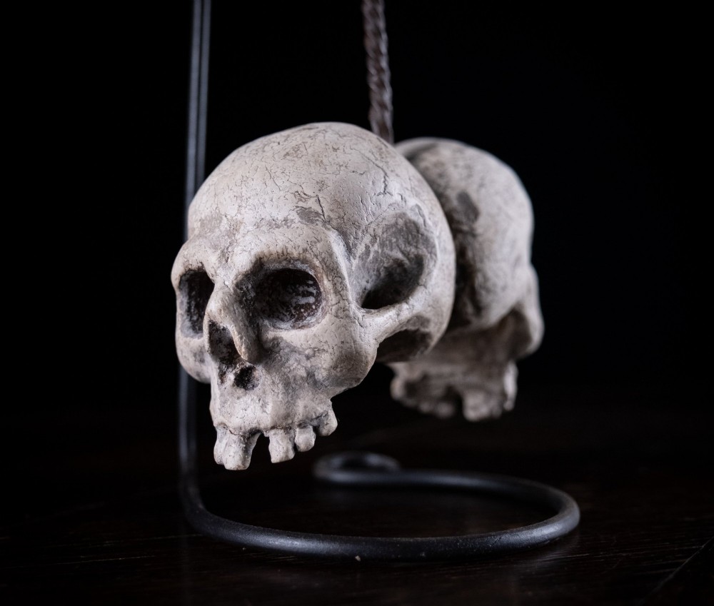 Two miniature carved skulls suspended by a metal chain from a black iron display stand. View II