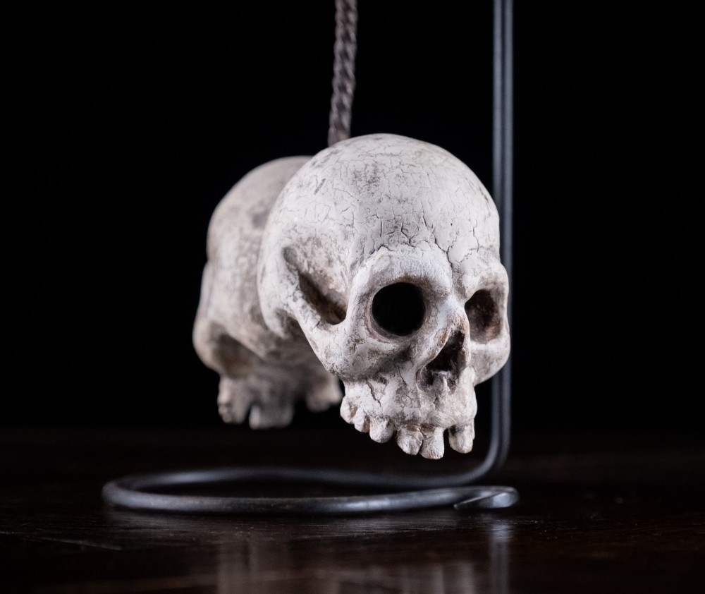 Two miniature carved skulls suspended by a metal chain from a black iron display stand. View III