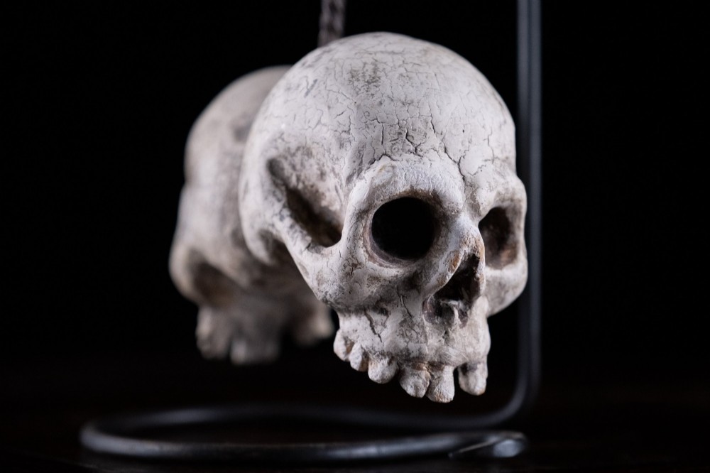 Two miniature carved skulls suspended by a metal chain from a black iron display stand. View V