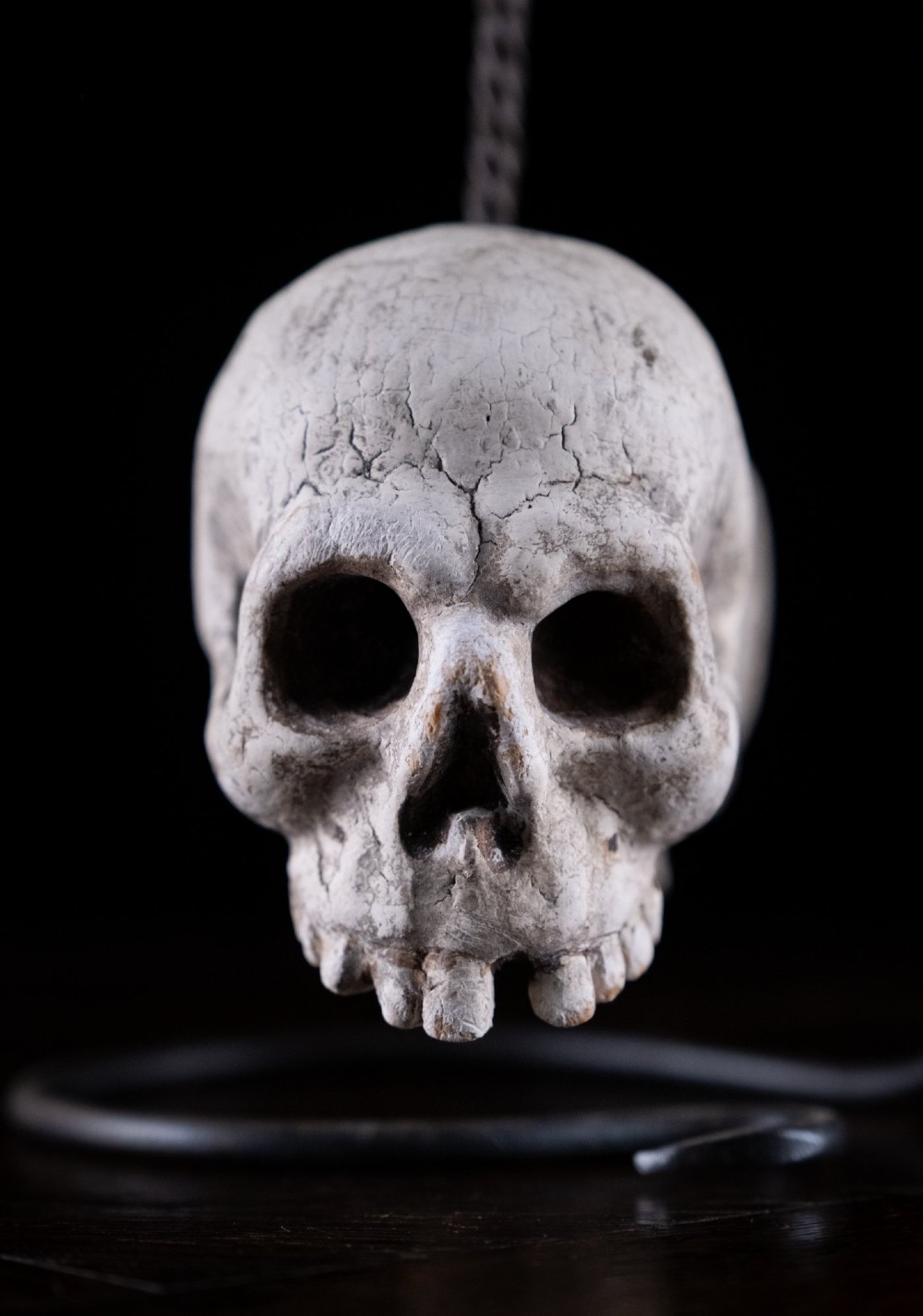 Two miniature carved skulls suspended by a metal chain from a black iron display stand. View VI