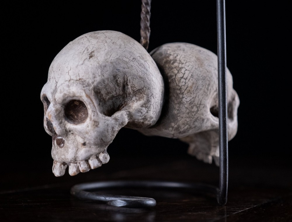 Two miniature carved skulls suspended by a metal chain from a black iron display stand. View IX