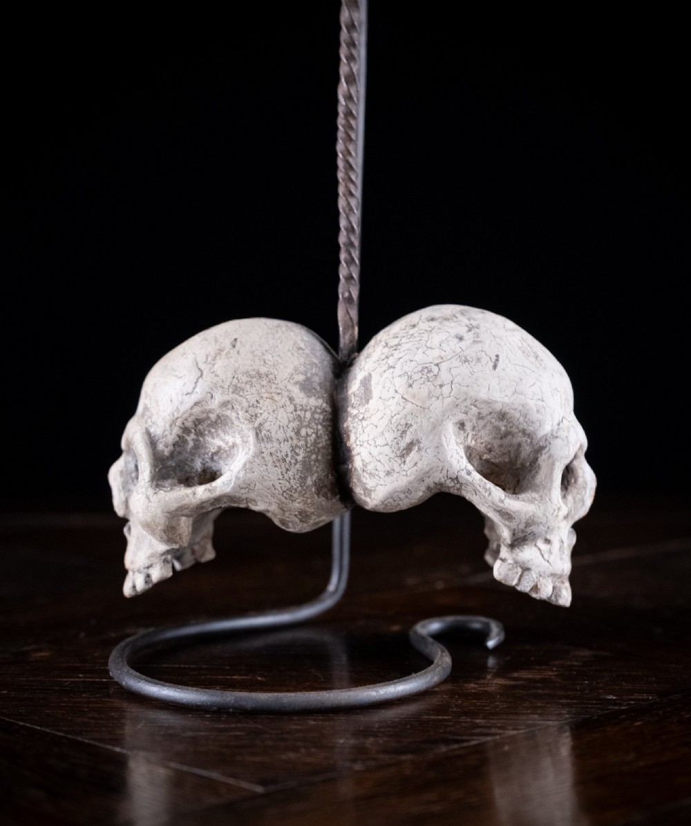 Two miniature carved skulls suspended by a metal chain from a black iron display stand. View X