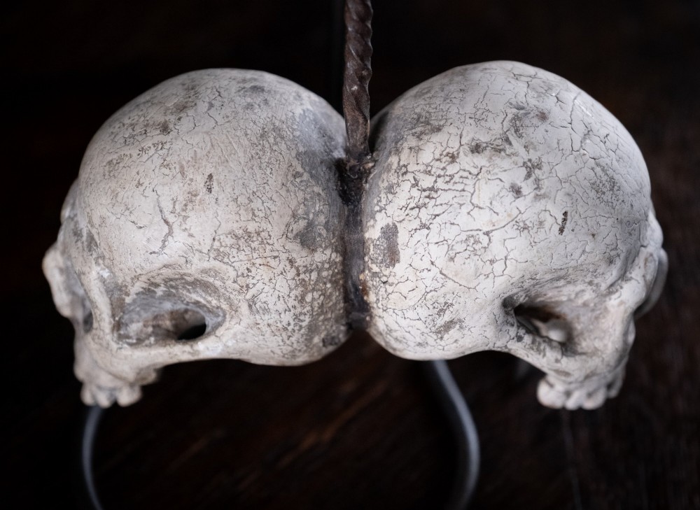 Two miniature carved skulls suspended by a metal chain from a black iron display stand. View XI