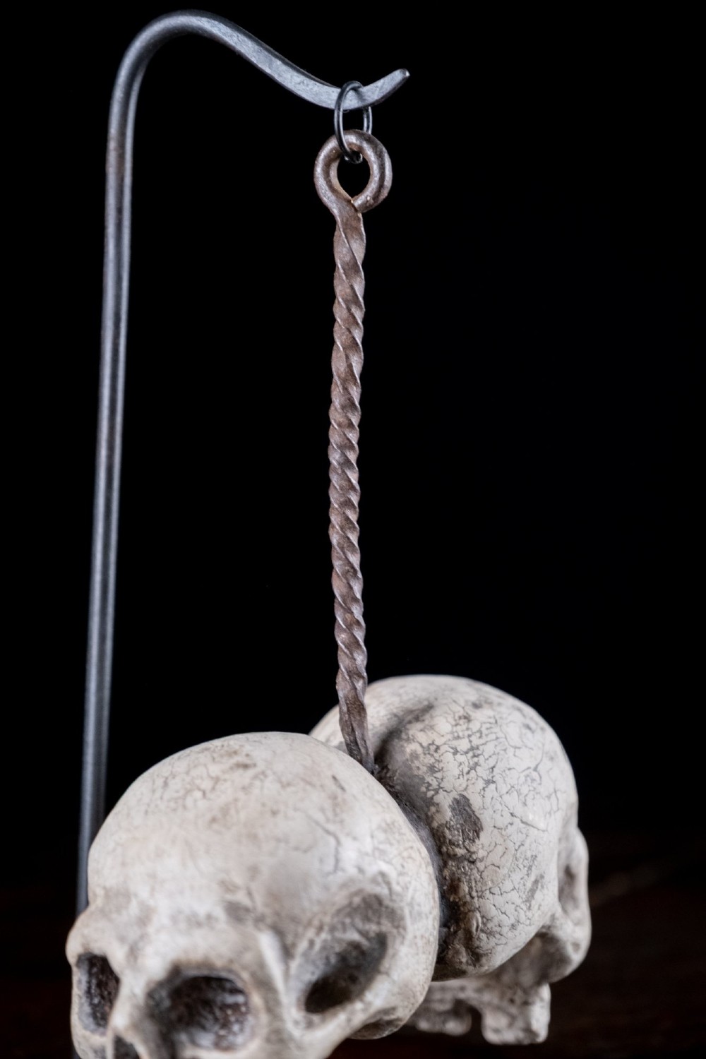 Two miniature carved skulls suspended by a metal chain from a black iron display stand. View XII