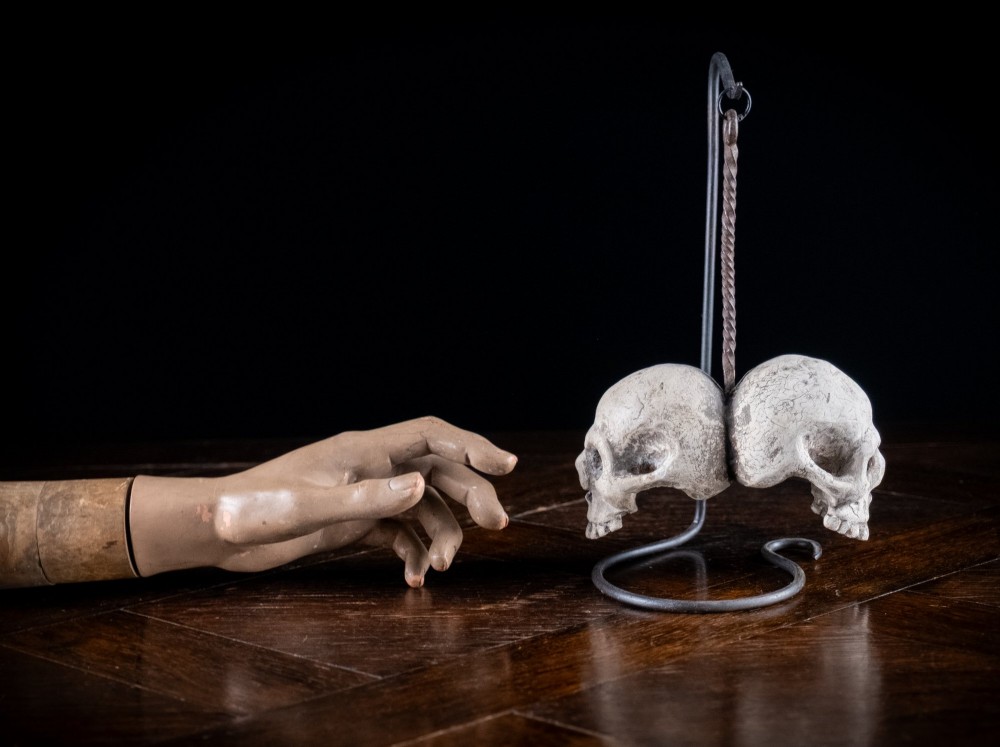 Two miniature carved skulls suspended by a metal chain from a black iron display stand. View XIV