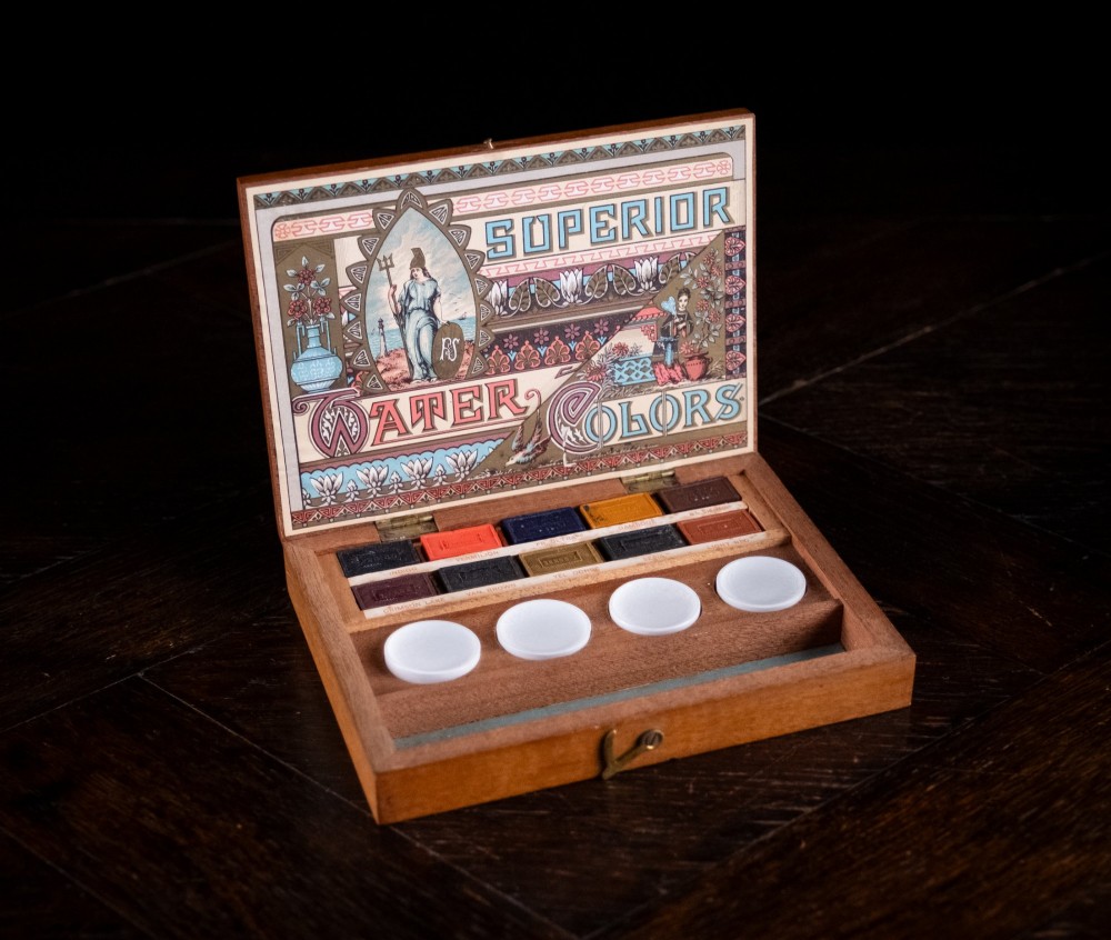 An open Victorian mahogany watercolour box displaying pigment cakes, four round porcelain mixing dishes, and an illustrated inner lid label reading Superior Water Colors.