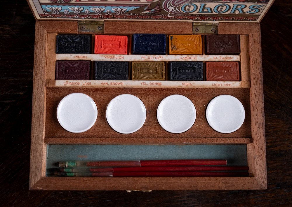 An open Victorian mahogany watercolour box displaying pigment cakes, four round porcelain mixing dishes, and an illustrated inner lid label reading Superior Water Colors. View II
