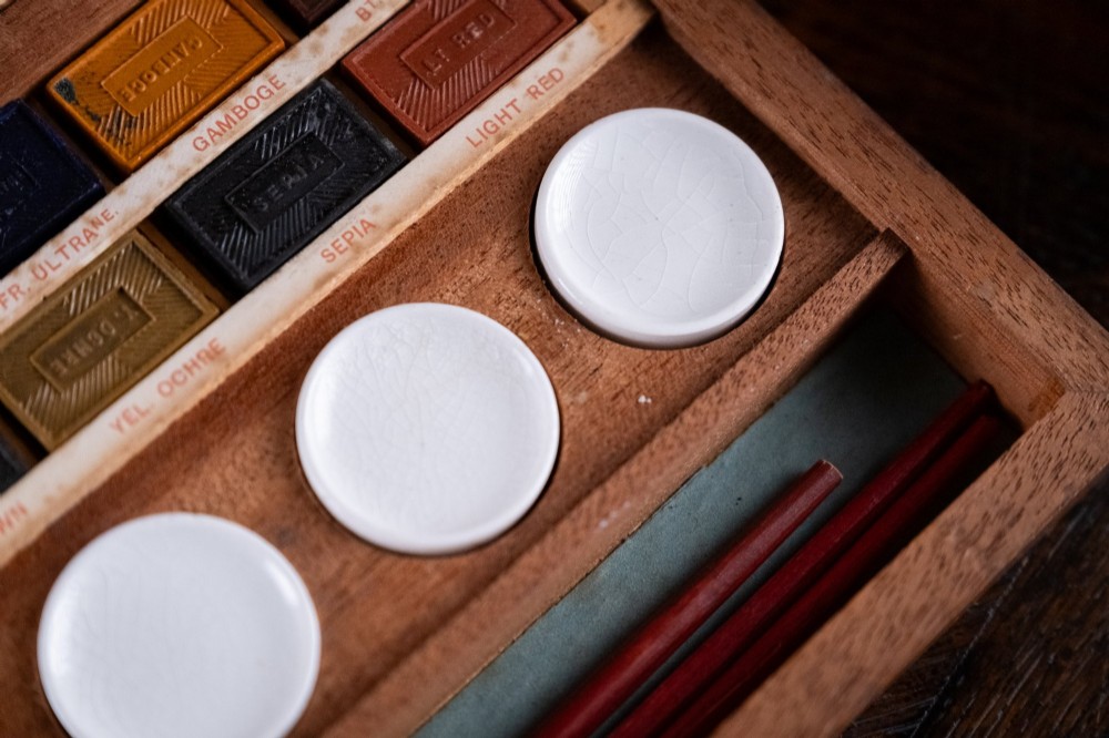 An open Victorian mahogany watercolour box displaying pigment cakes, four round porcelain mixing dishes, and an illustrated inner lid label reading Superior Water Colors. View III