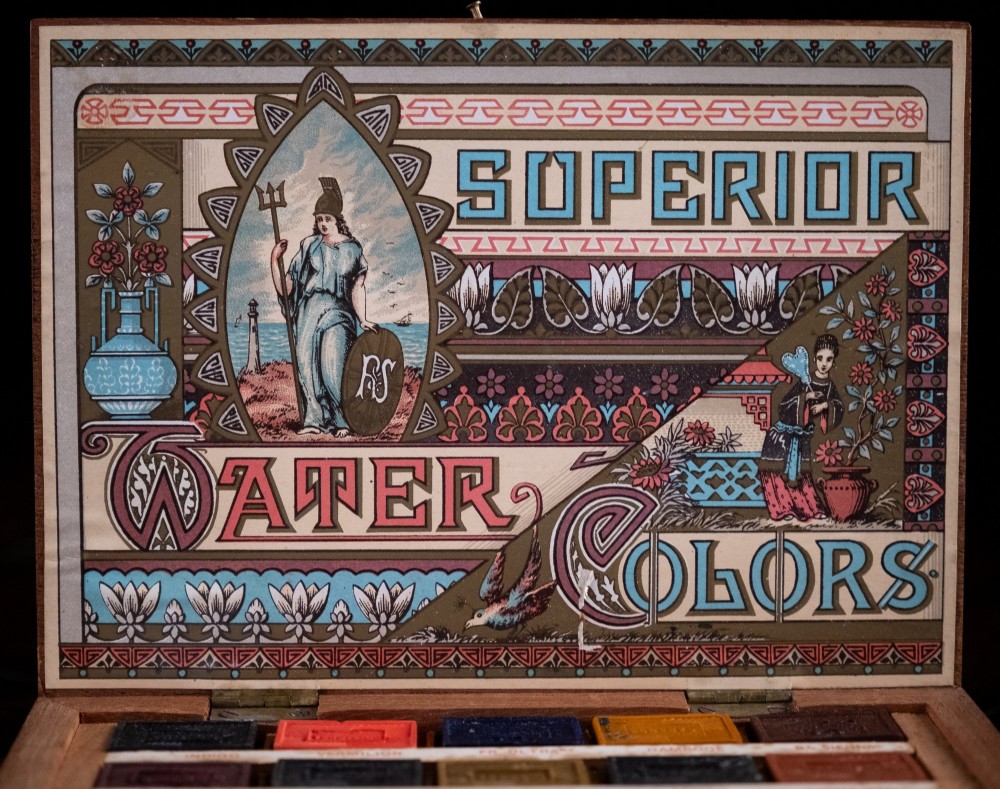An open Victorian mahogany watercolour box displaying pigment cakes, four round porcelain mixing dishes, and an illustrated inner lid label reading Superior Water Colors. View IV