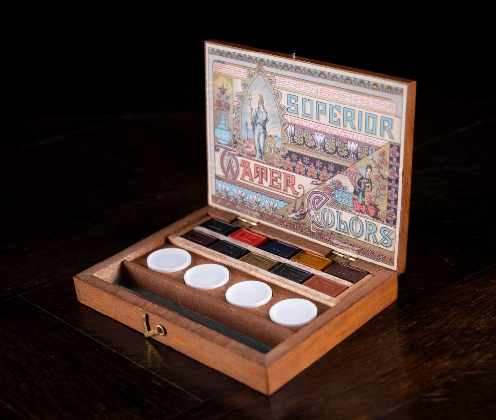 An open Victorian mahogany watercolour box displaying pigment cakes, four round porcelain mixing dishes, and an illustrated inner lid label reading Superior Water Colors. View V