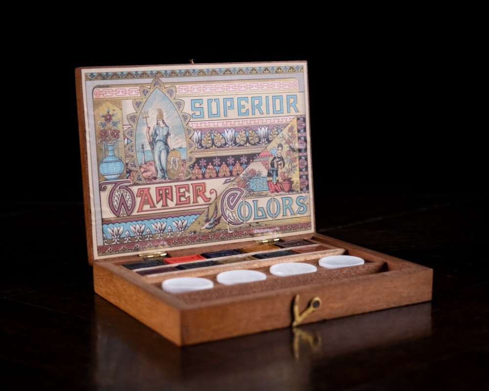 An open Victorian mahogany watercolour box displaying pigment cakes, four round porcelain mixing dishes, and an illustrated inner lid label reading Superior Water Colors. View VI