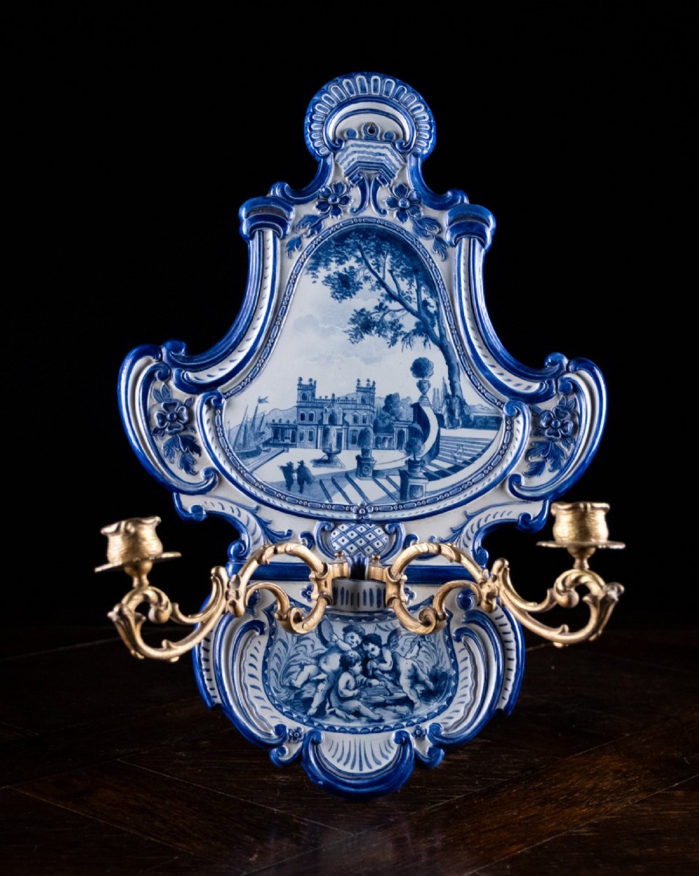 A late nineteenth-century blue and white ceramic wall sconce featuring Rococo scrollwork framing painted architectural and cherub motifs, fitted with two gilt bronze candle arms.