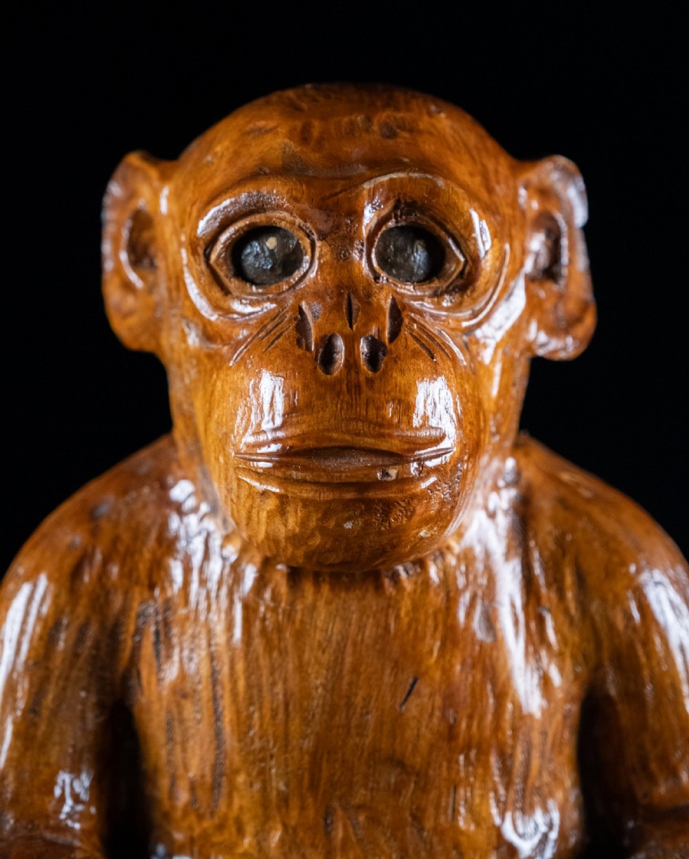 A hand-carved warm brown wooden sculpture depicting a seated monkey with chiselled fur texture. View X
