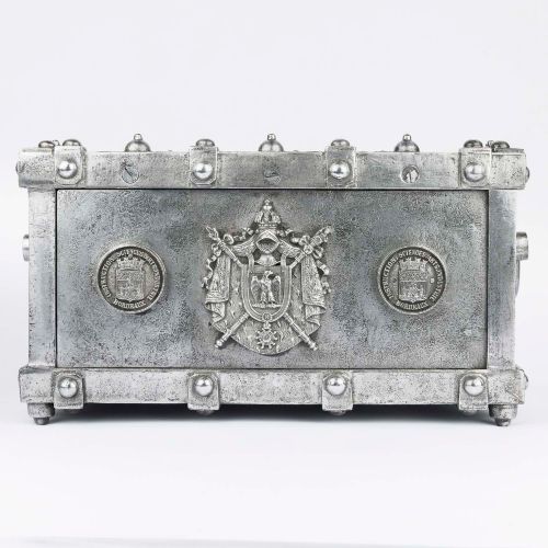 fig 1. A heavy rectangular metal strongbox featuring riveted steel straps, raised studs, an embossed coat of arms, and maker plaques.