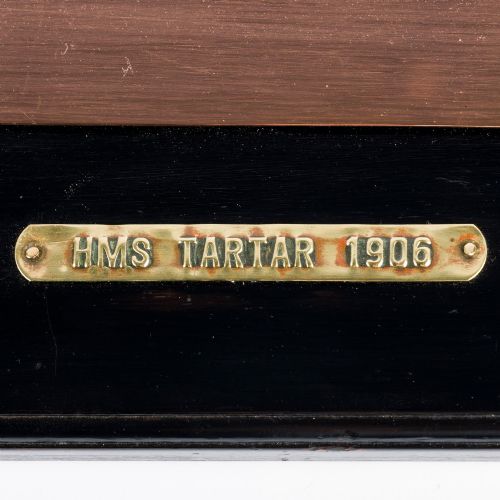 fig 1. A rectangular black lacquered wooden plaque displaying a copper cutout profile of HMS Tartar 1906 with a small brass title tag below.