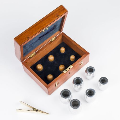 fig 1. An open mahogany wooden box lined with blue velvet, containing six rounded metal egg weights and brass tweezers fitted into the lid.
