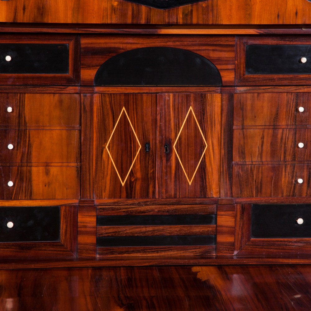 A tall Biedermeier secretaire a abattant featuring rich bookmatched brown wood veneers, an architectural pediment top, a fall-front desk section, and lower drawers with ebonised detail. View VII
