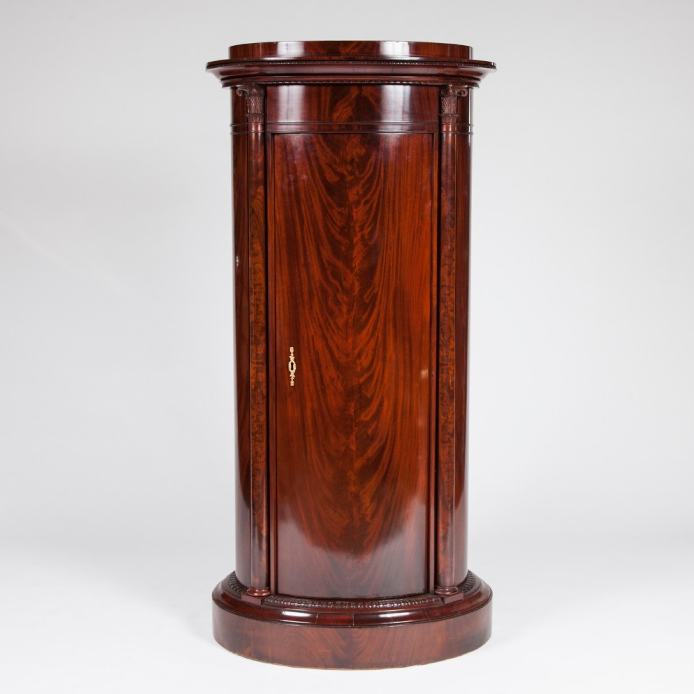 An antique Danish oval pedestal cabinet in polished flame mahogany featuring a single curved door, carved pilaster columns, and a brass keyhole escutcheon.