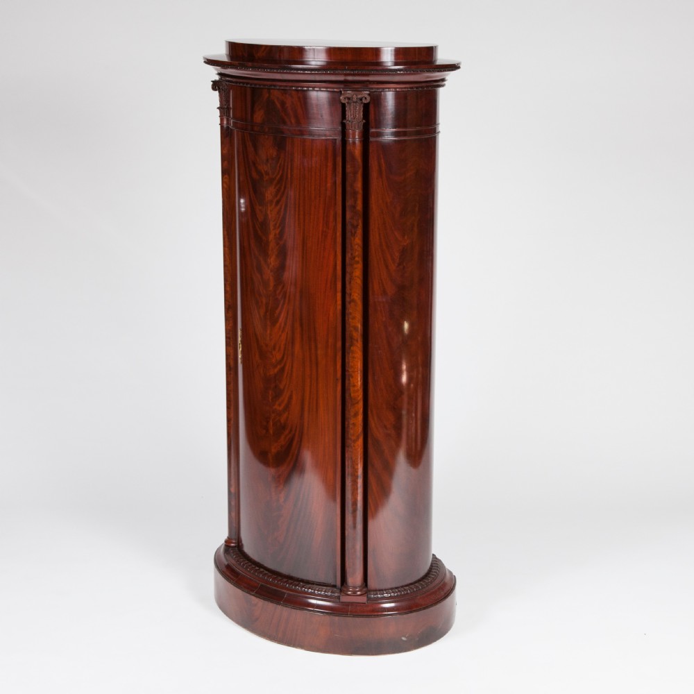 An antique Danish oval pedestal cabinet in polished flame mahogany featuring a single curved door, carved pilaster columns, and a brass keyhole escutcheon. View IV