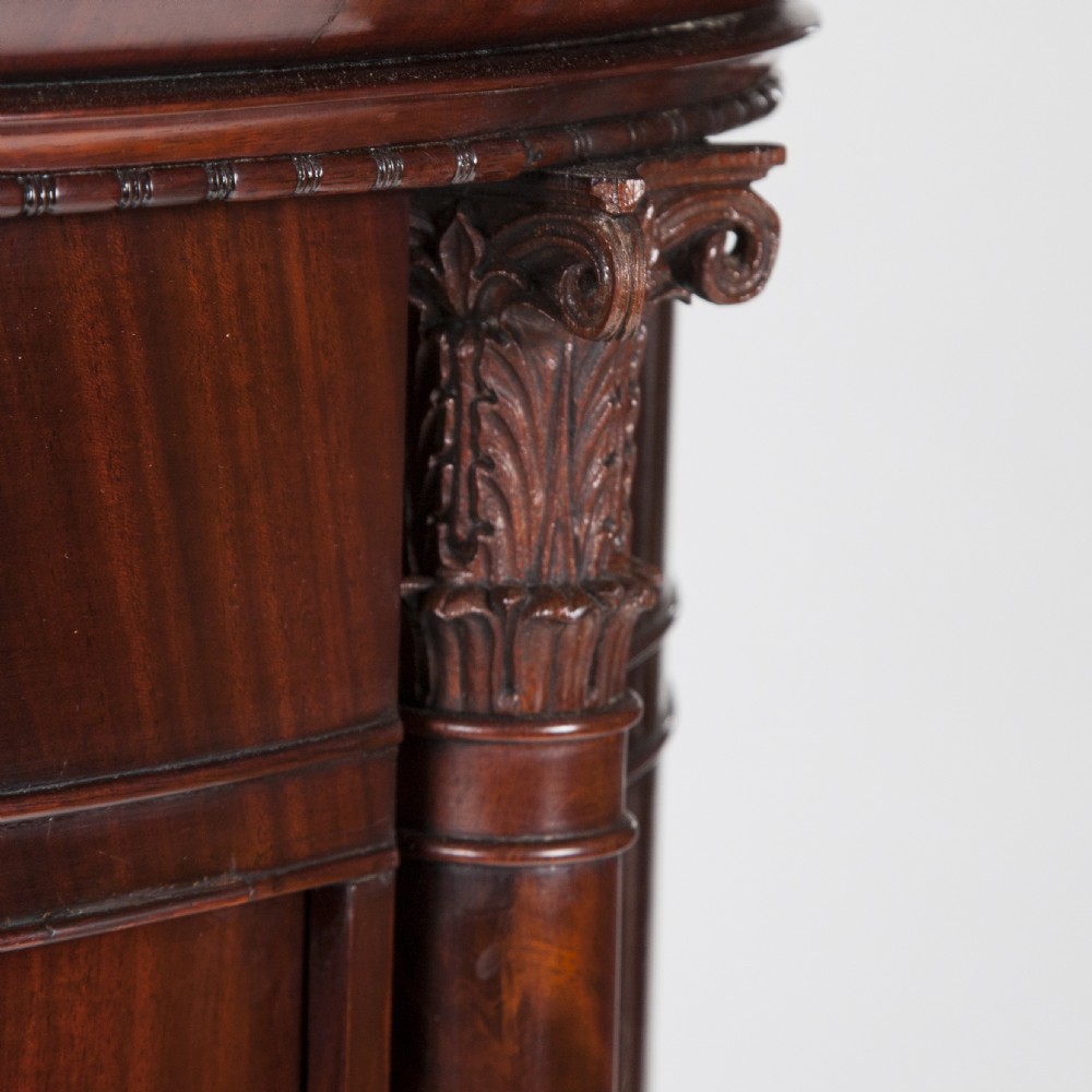 An antique Danish oval pedestal cabinet in polished flame mahogany featuring a single curved door, carved pilaster columns, and a brass keyhole escutcheon. View VI
