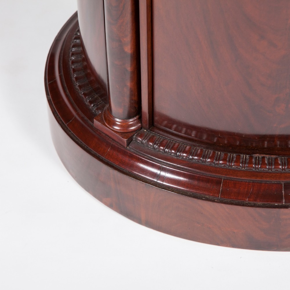 An antique Danish oval pedestal cabinet in polished flame mahogany featuring a single curved door, carved pilaster columns, and a brass keyhole escutcheon. View VIII